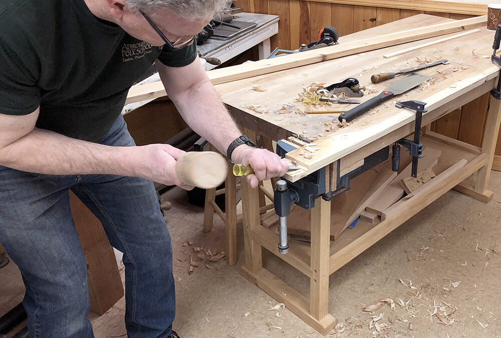 A man leans over a work bench with a wooden mallet in hand, chiseling a piece of wood.